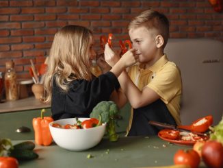 Playful boy and girl wearing apron preparing healthy vegetarian salad together at table with ingredients and bowls while looking at each other through slices of pepper