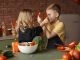 Playful boy and girl wearing apron preparing healthy vegetarian salad together at table with ingredients and bowls while looking at each other through slices of pepper